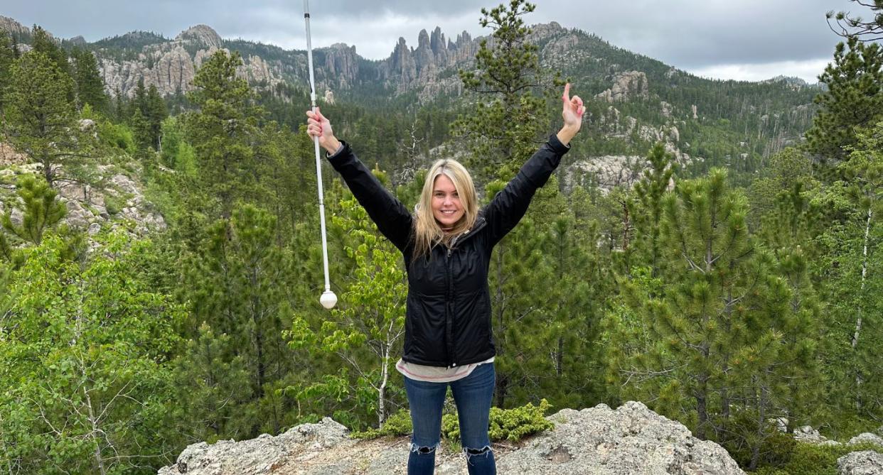 Claire Sisk posing on top of a mountain with greenery behind her, holding her cane up high both arms in the air. (Supplied)