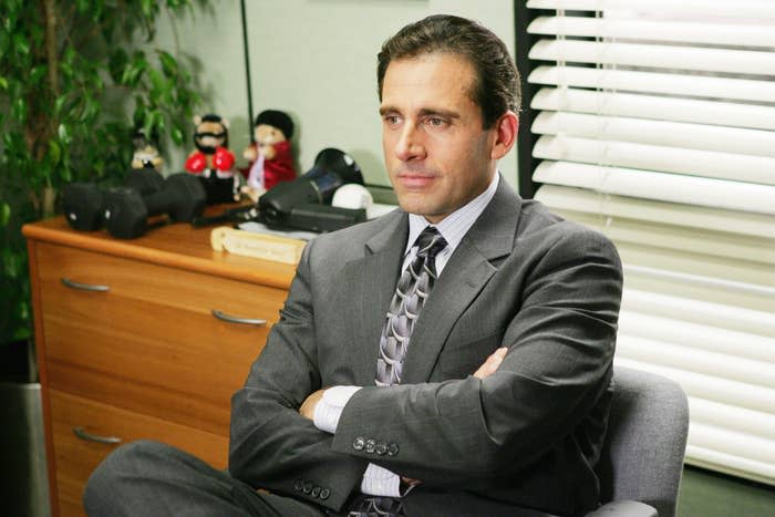 Michael Scott, dressed in a suit and tie, sits with arms crossed at a desk with office items and blinds in the background