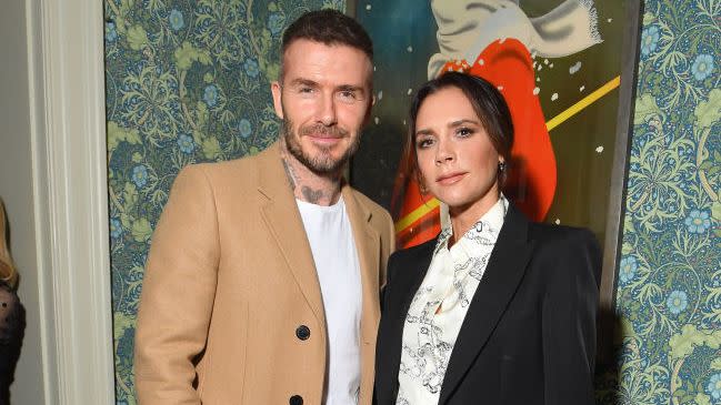 david beckham and victoria beckham stand and pose for a photo together, he wears a tan coat with a white shirt and dark pants, she wears a black suit with a white patterned shirt, they stand in front of a piece of artwork hanging on a wall with blue and green floral wallpaper