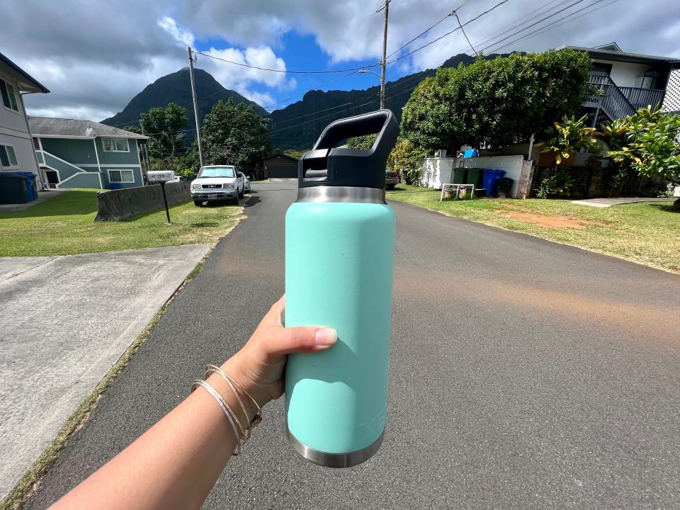 The writer holds a blue reusable water bottle in front of a street and mountains in Hawaii
