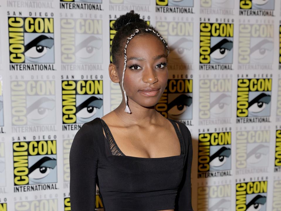 kyo ra on the san diego comic con red carpet, wearing a long-sleeve black drop top and high waisted jeans, belted with a colorful scarf. her hair is tied up in a fun, with two contrasting white braids framing her face.