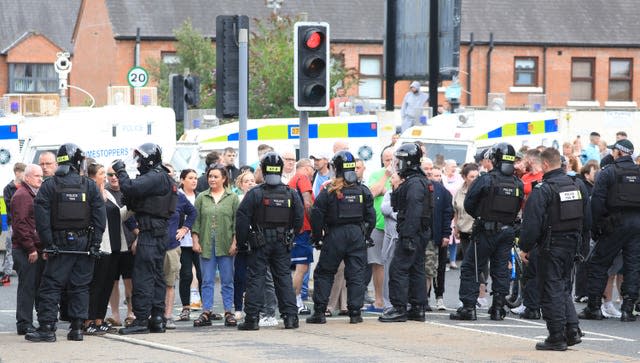 A line of riot police stand in front of a large group of people on a street