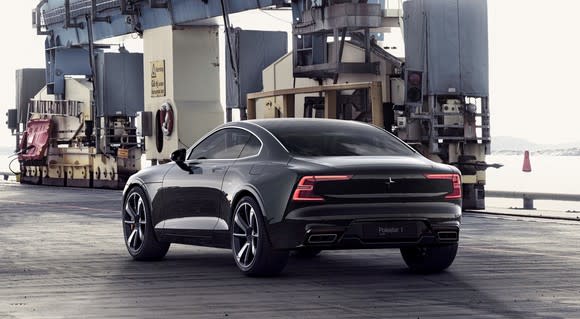 A black Polestar 1 coupe near a waterfront, with industrial equipment in the background.