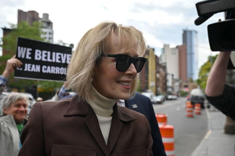 E. Jean Carroll smiling on a city street, wearing sunglasses and a brown coat; supporters in the background hold a "WE BELIEVE JEAN CARROLL" sign