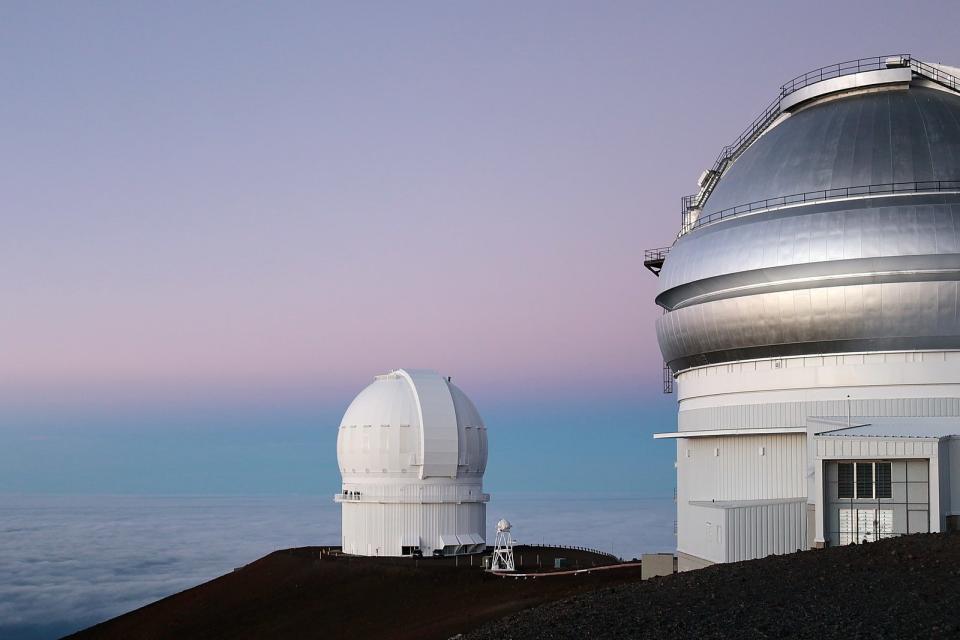 Two telescopes in Hawaii look up at a bluish-pink sky, scanning a wide-field view of the solar system.