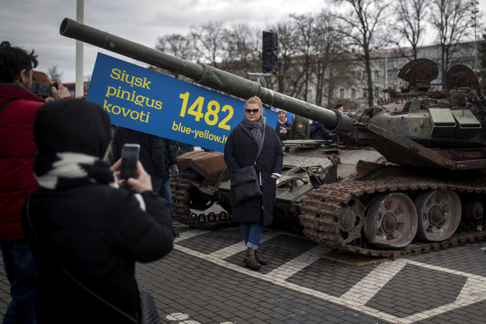 A woman poses for a photo in front of the destroyed Russian tank T-72B installed as a symbol of war marking the first anniversary of Russia's full-scale invasion of Ukraine, decorated with the banner reading "Send money to fight", at Cathedral Square in Vilnius, Lithuania, Wednesday, March 1, 2023. Some ethnic Russians in the Baltic states have placed flowers at displays of burnt-out Russian tanks seized by Ukrainians, making a gesture of homage and support for Russia's war against Ukraine. (AP Photo/Mindaugas Kulbis)