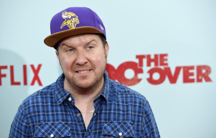 A white man with facial hair, a purple hat and a blue plaid shirt smiling and looking to his side in front of a blue backdrop