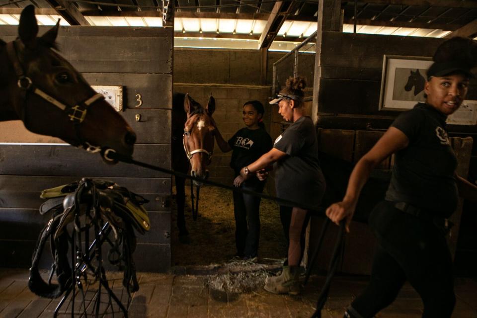 A woman leading a horse to its stall as others in the stable work with another horse