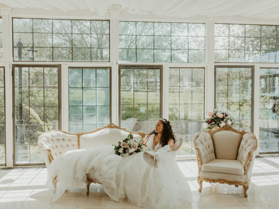 A bride sits on a couch in front of a wall of windows.