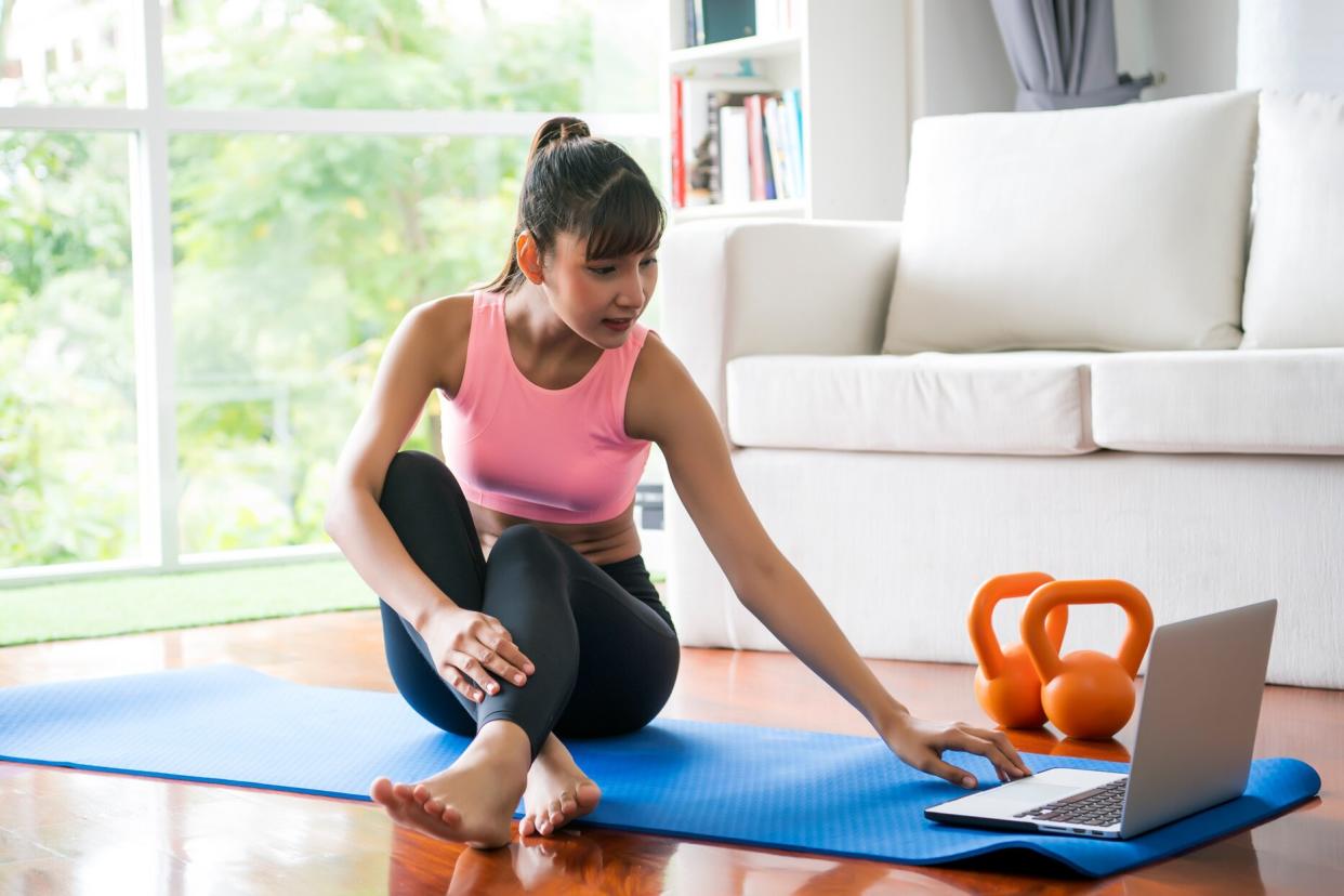 person sitting on yoga mat on living room floor looking at laptop with kettlebells nearby