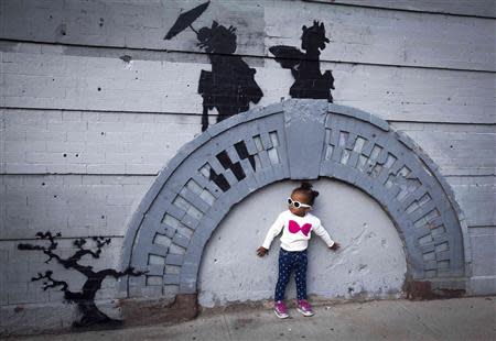 A child poses for a photo under a new art piece by British graffiti artist Banksy in the Brooklyn borough of New York, October 17, 2013. REUTERS/Carlo Allegri