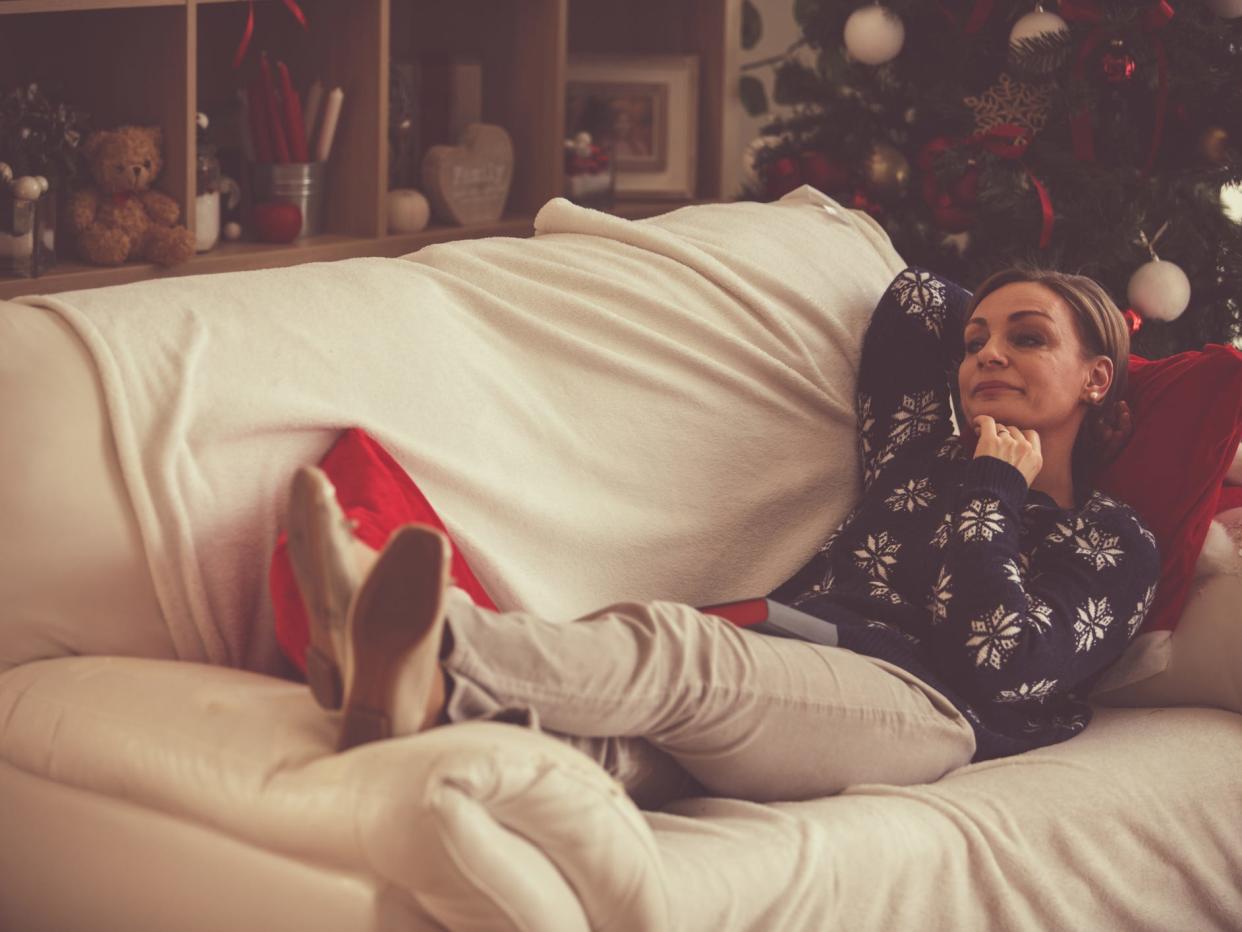 A woman with light blonde hair tied back sits horizontally on a white leather couch with her feet up on the arm. The couch is covered with a white blanket. The woman wears a blue knit sweater with white snowflakes, off-white pants, and white flats. She has one arm tucked behind her head and the other is bent, with her hand in a fist under her chin with a thoughtful expression on her face. Behind her is a decorated Christmas tree and shelves with photos and random objects.