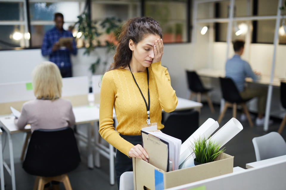 Stressed young woman in yellow sweater standing at table and touching face with hand while packing stuff in office after dismissal