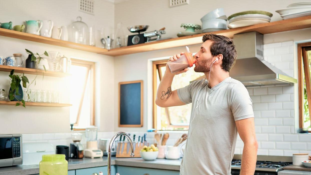 one fit young caucasian man drinking bottle of chocolate whey protein shake for energy for training workout while wearing earphones in a kitchen at home guy having sports supplement for muscle gain and dieting with weightloss meal replacement