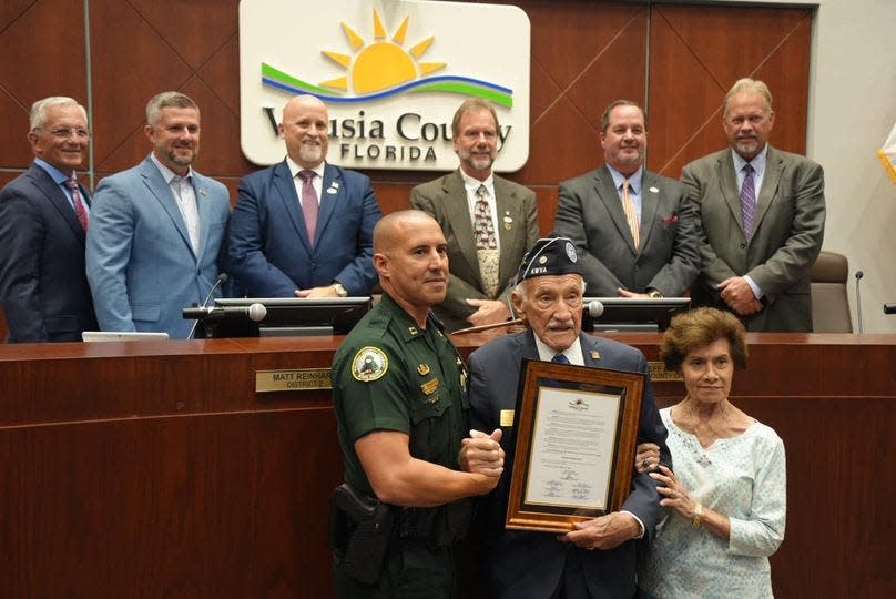 Korean War veteran Joseph Sicinski poses with his wife, Anita, and grandson Capt. Kurt Schoeps of the Volusia Sheriff's Office while holding a proclamation that named May 21, 2024, as "Joseph Sicinski Day." Members of the Volusia County Council stand on the dais.