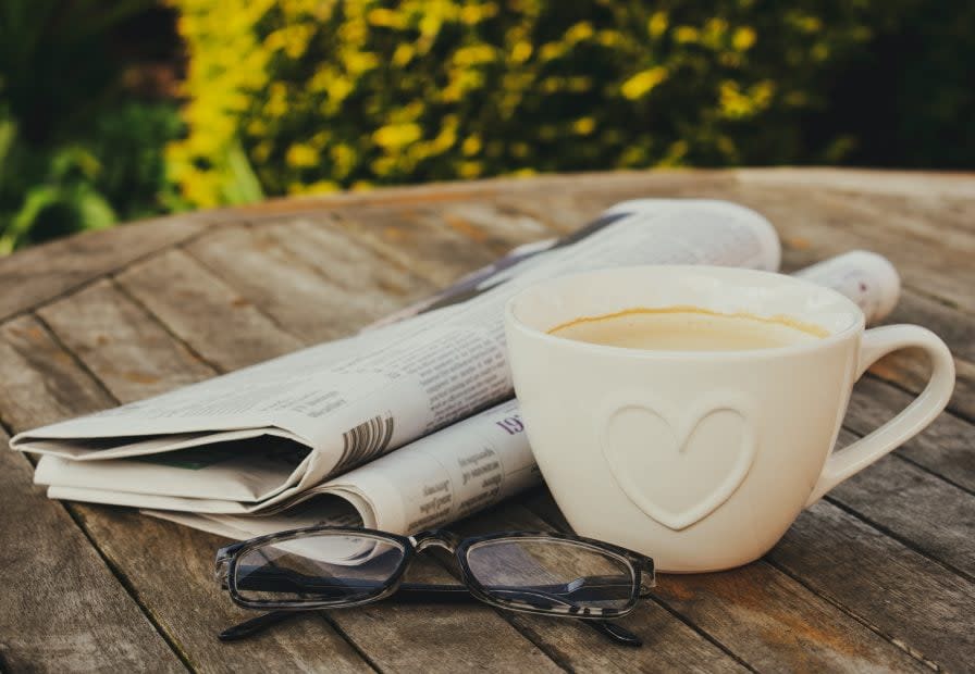 Eine Zeitung, eine Kaffeetasse und eine Brille stehen auf einem Holztisch im Garten.