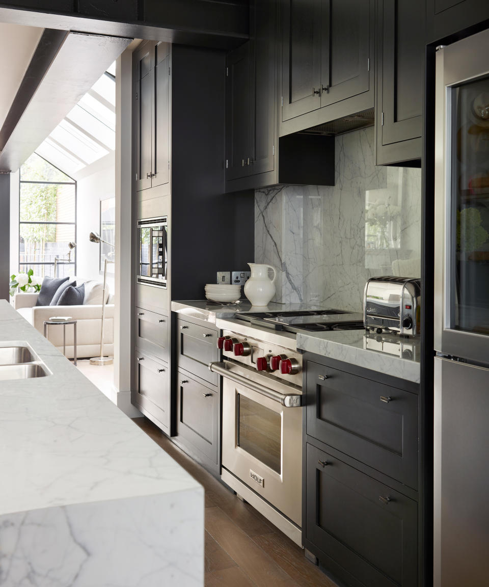 A kitchen with dark blue cabinets next to a built-in range cooker and a marble effect island with a view into a living space with a glass roof