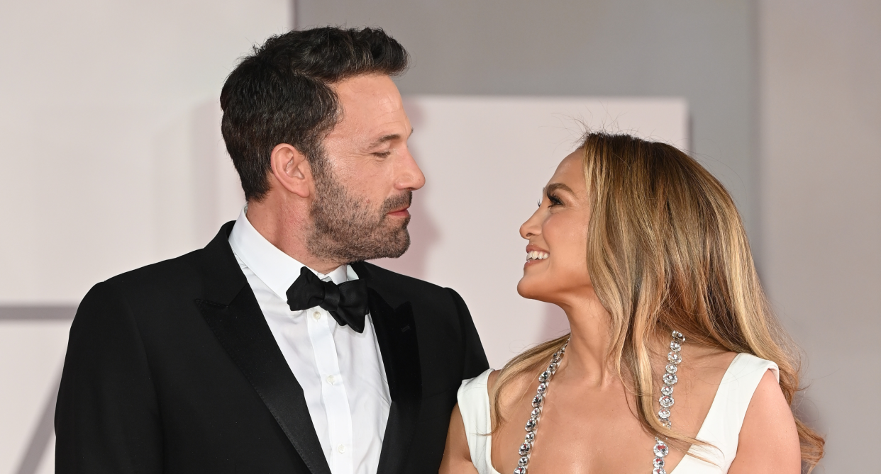 jennifer lopez and ben affleck looking at each other on red carpet, j.lo in white dress and ben affleck in black suit tuxedo jacket