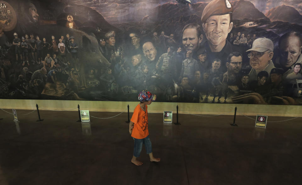 In this Saturday, June 22, 2019, photo, a boy watches a painting depicting the recuse mission to save 12 soccer boys and their coach near the Tham Luang cave in Mae Sai, Chiang Rai province, Thailand. Some of the 12 young Thai soccer players and their coach have marked the anniversary of their ordeal that saw them trapped in a flooded cave for two weeks with a commemorative marathon in northern Thailand. (AP Photo/Sakchai Lalit)