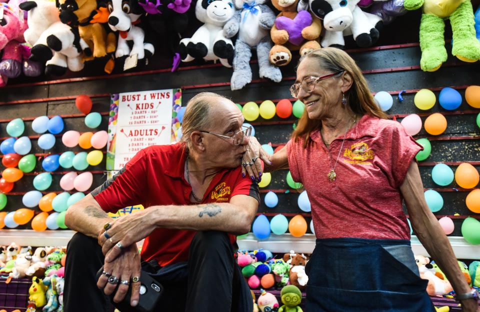 Stephen Stimpson of Bangor, Maine, kisses his wife Tina's hand Friday, July 16, 2022, in their game booth at the Eaton County Fair in Charlotte after Tina spoke of how they met 42 years ago working a carnival.  He offered to smoke a joint with me and halfway through he blew me a shotgun and our lips touched and that was it. We both saw fireworks and we've been together ever since."