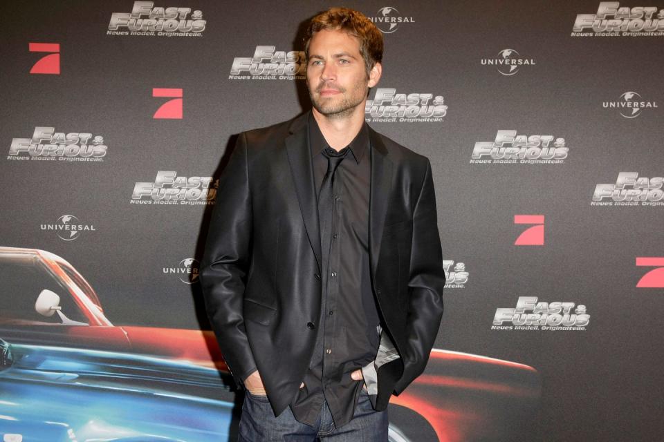 paul walker wearing a black leather coat and black shirt, standing in front of a fast and furious display wall