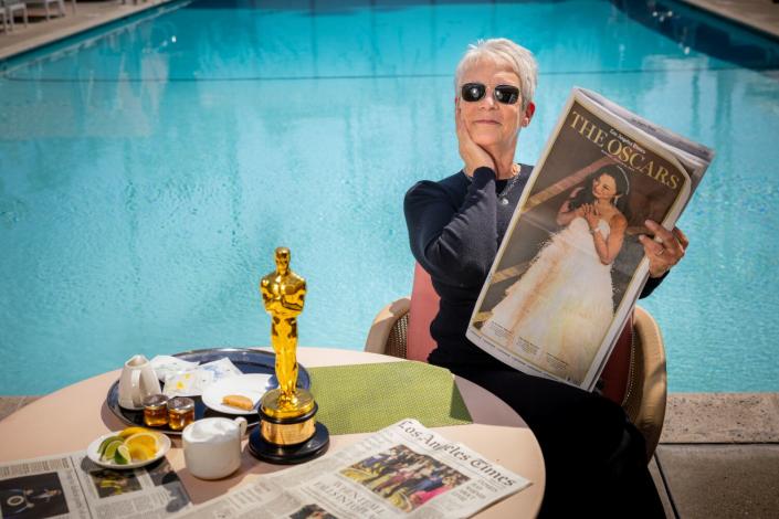 A woman in sunglasses holds a newspaper section next to a table with her Oscar on it.