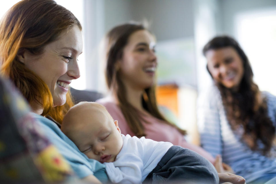 Smiling mother with sleeping son sitting by friends at home. Happy females are with baby boy in domestic room. They are spending leisure time.