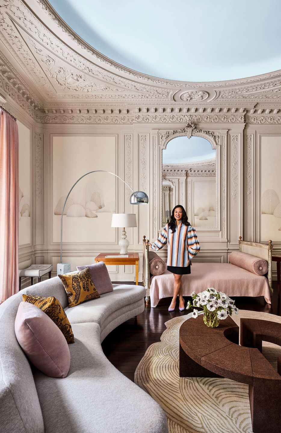 a woman with long dark hair wearing a striped top and black skirt stands next to a daybed, curved gray sofa, amorphous rug with lines, two half moon cocktail tables, end table with lamp, curved floor lamp with silver globe