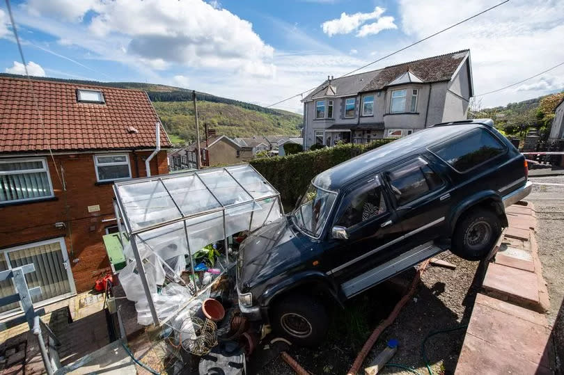 Black 4x4 vehicle perched at top of garden, its front having crashed into a greenhouse