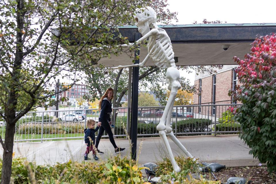 Hank R., 2, walks with his nanny Allison Alderton by a giant skeleton at the entrance of Royal Oak Public Library in Royal Oak on Wednesday, Oct. 25, 2023.