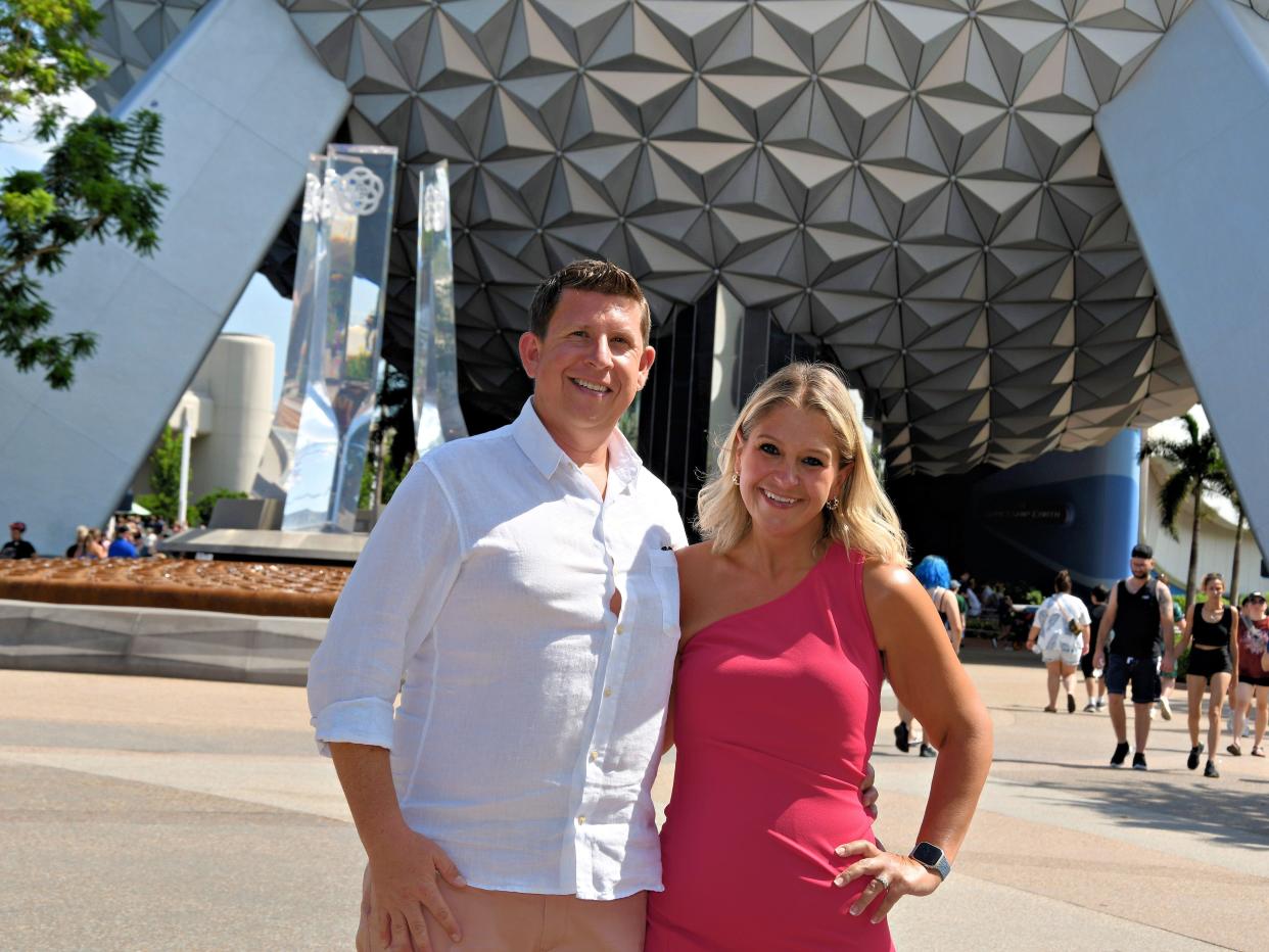 Terri Peters and her husband standing in front of the Spaceship Earth sculpture at Walt Disney's EPCOT. Terri stands on the right with her hand on her waist and her other hand around her husband. She has dyed blonde hair, dark brown eyes, and wears a hot pink one shouldered dress. Her husband has brown hair that's flipped up at the front and wears a white buttondown shirt and khaki pants. His left hand is around Terri. They smile widely.