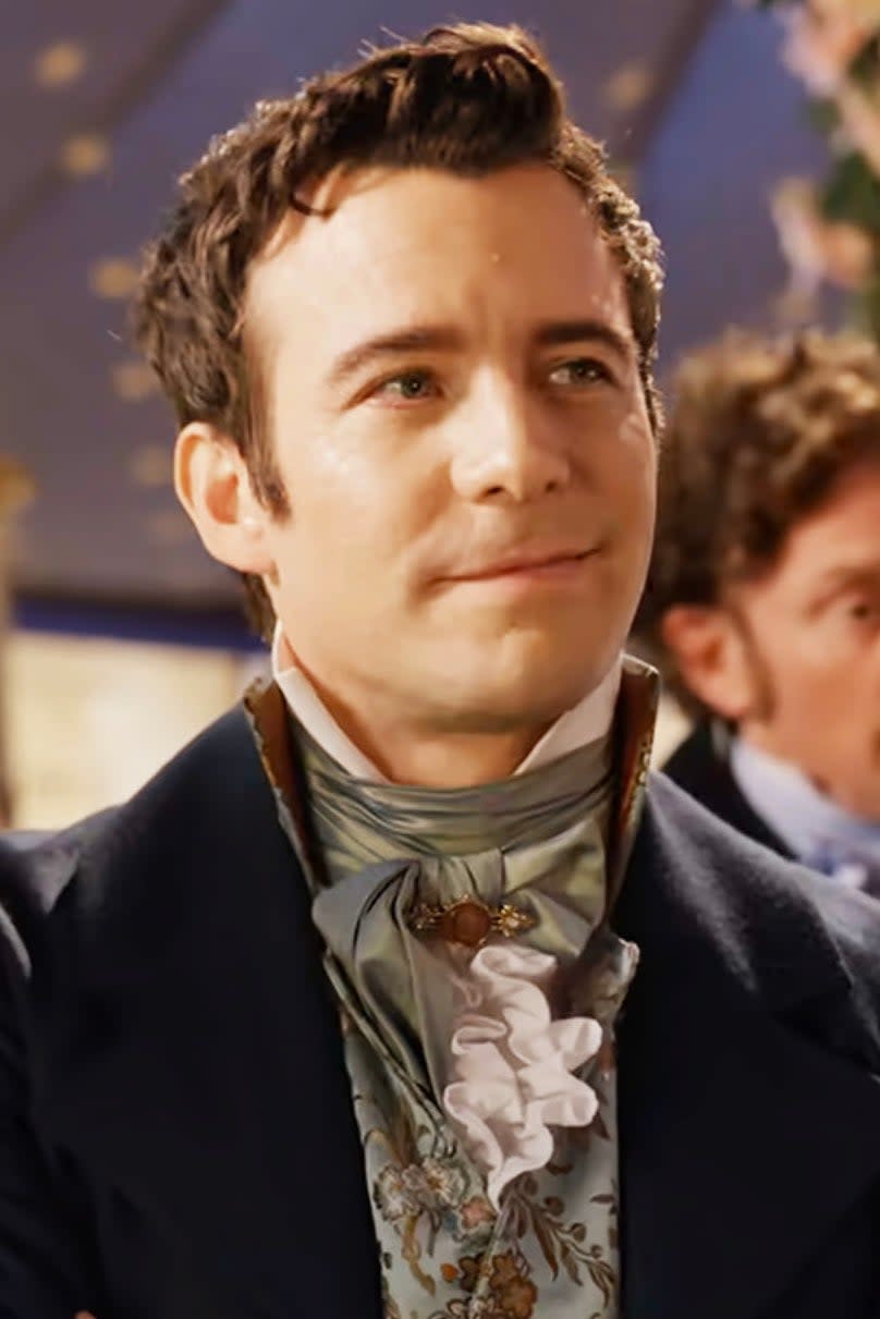 Luke Thompson in 'Bridgerton', wearing a Regency-style suit with a decorative cravat, stands confidently with arms crossed at a social event
