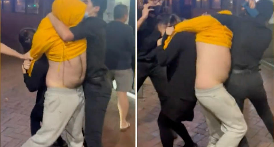 Man in yellow jumper being held back by restaurant staff in black