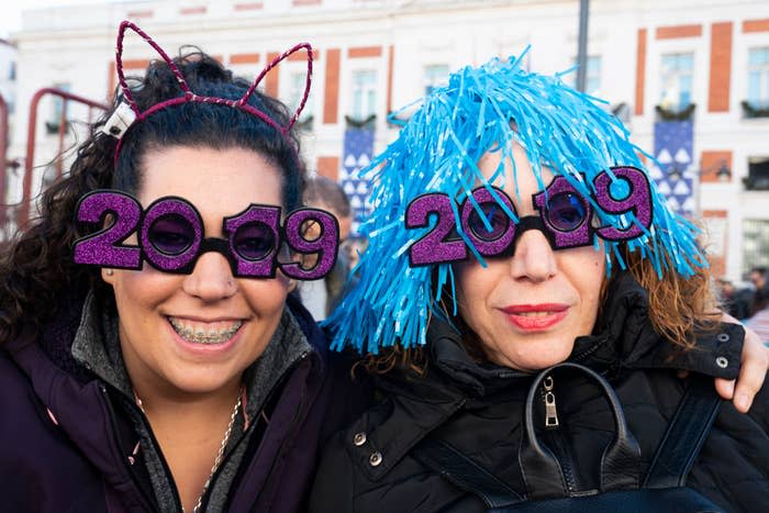 Two women wearing "2019" glasses, one with cat ear headband and the other with a blue wig, celebrate New Year's Eve outdoor