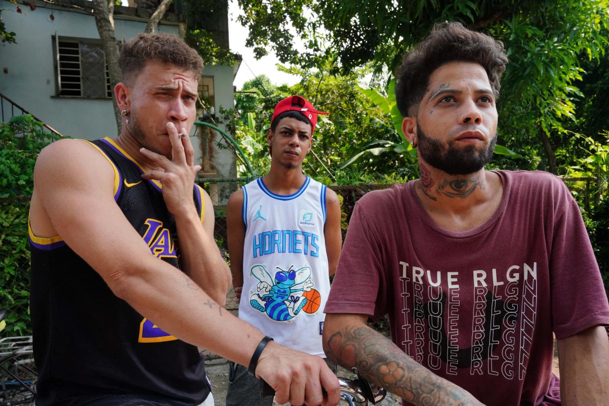 three young men, two in NBA jerseys, one one left smoking cigarette, one in middle wearing backwards red cap, one on right with tattoos on neck and arms