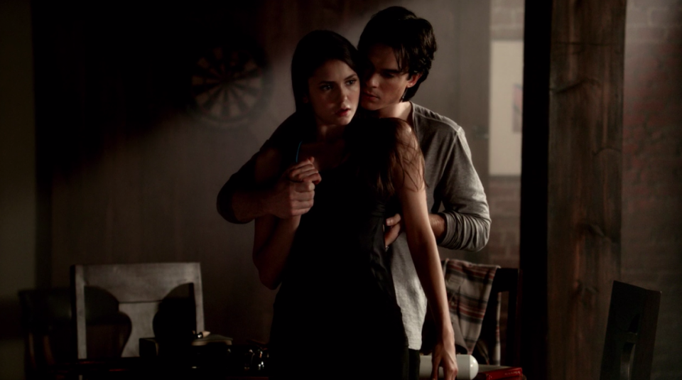 "The Vampire Diaries" characters Damon and Elena stand in front of each other, as he touches her back and hand