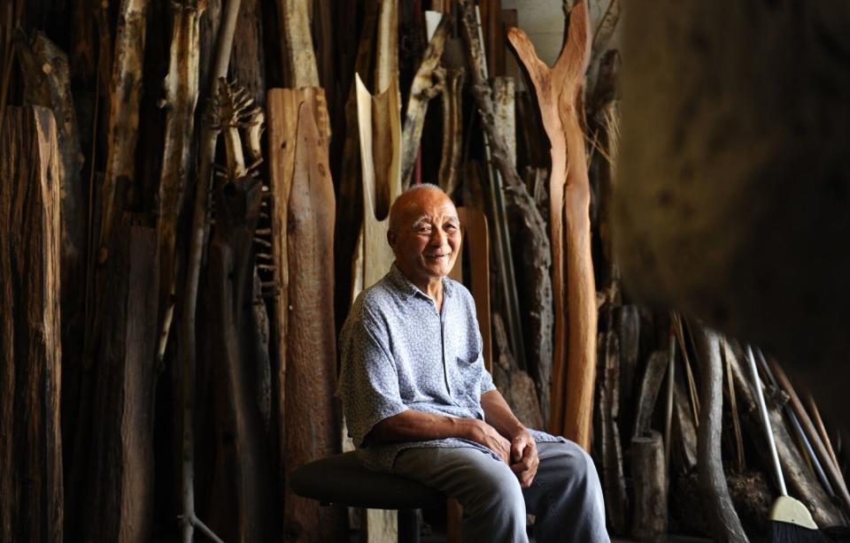 Kenzi Shiokova smiles as he sits before rows of tree trunks in various states of sculptural fabrication