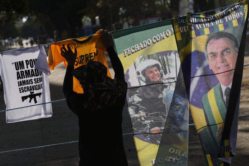 FILE - A vendor hangs electoral merchandise featuring Brazil's President Jair Bolsonaro, who is running for re-election, for sale during a campaign rally after a military parade commemorating the bicentennial of the country's independence in Brasilia, Brazil, Sept. 7, 2022. The shirt at left reads in Portuguese: "Armed people will never be enslaved." Supporters of incumbent President Jair Bolsonaro who refuse to accept his narrow defeat in October’s election have blocked roads and camped outside military buildings while pleading for intervention from the armed forces or marching orders from their commander in chief. (AP Photo/Eraldo Peres, File)