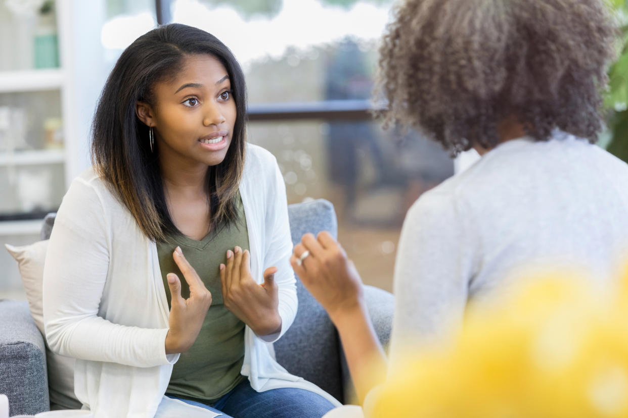 A teenage girl wears an intense expression as she gestures and argues with her unrecognizable mother on a couch in her living room.