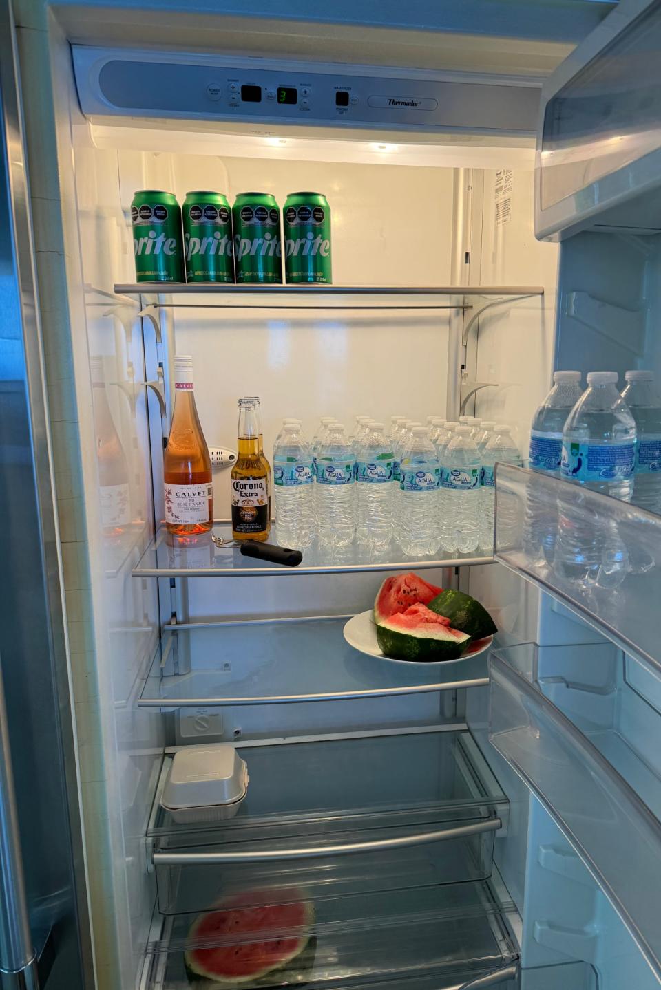 An open fridge contains several beverages and a plate with a cut watermelon. Drinks include Sprite, Clamato, Corona, Modelo, and bottles of water