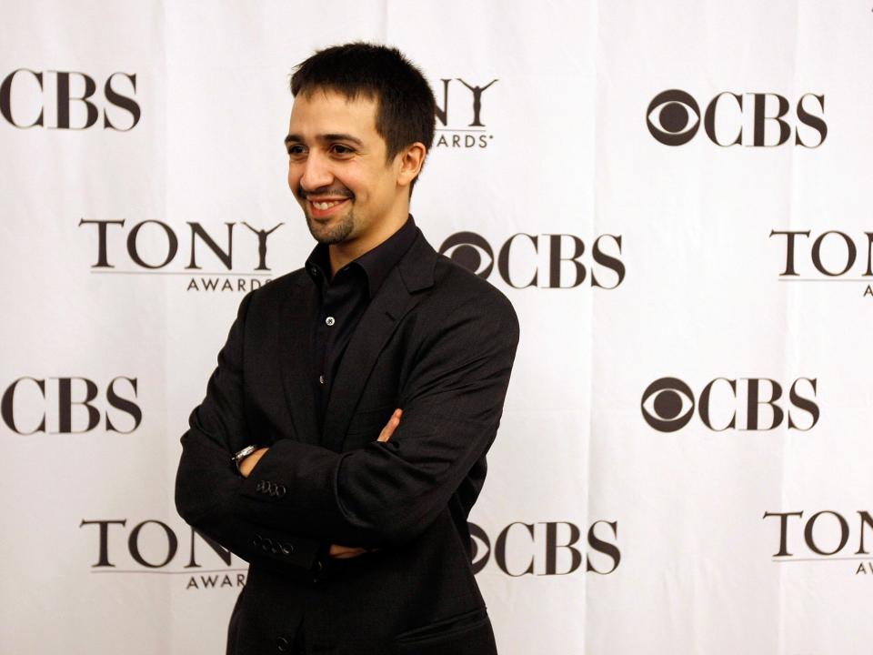 Lin-Manuel Miranda smiling and crossing his arms in front of a CBS Tony Awards backdrop.