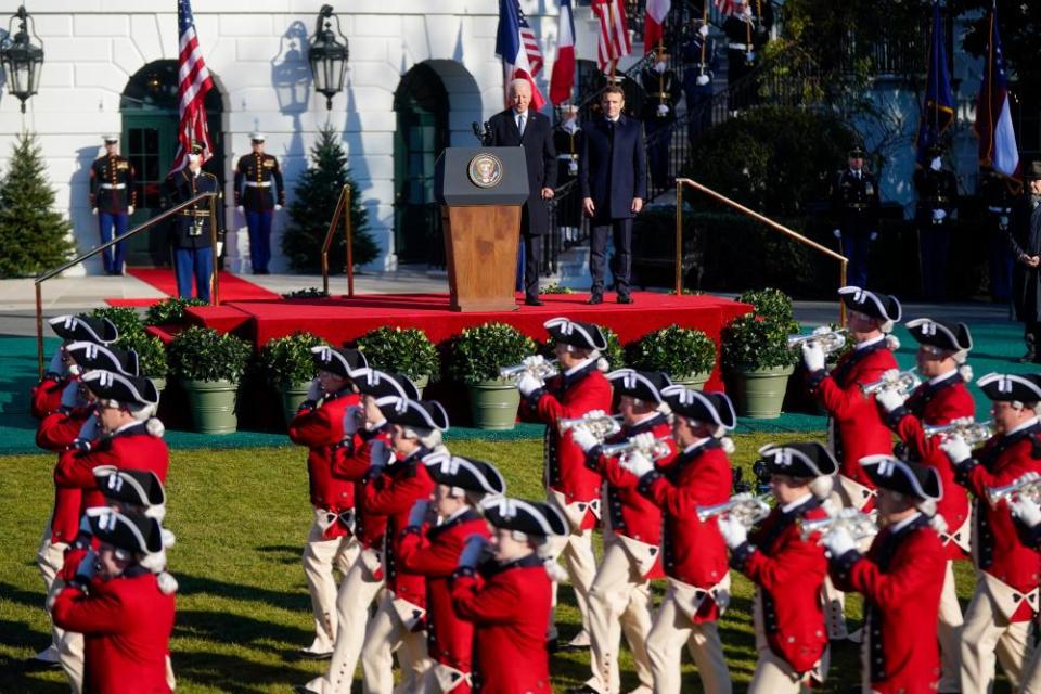 Soldiers dressed in red coats and 18th century military uniforms parade in front of a stage where Joe Biden and Emmanuel Macron are standing.