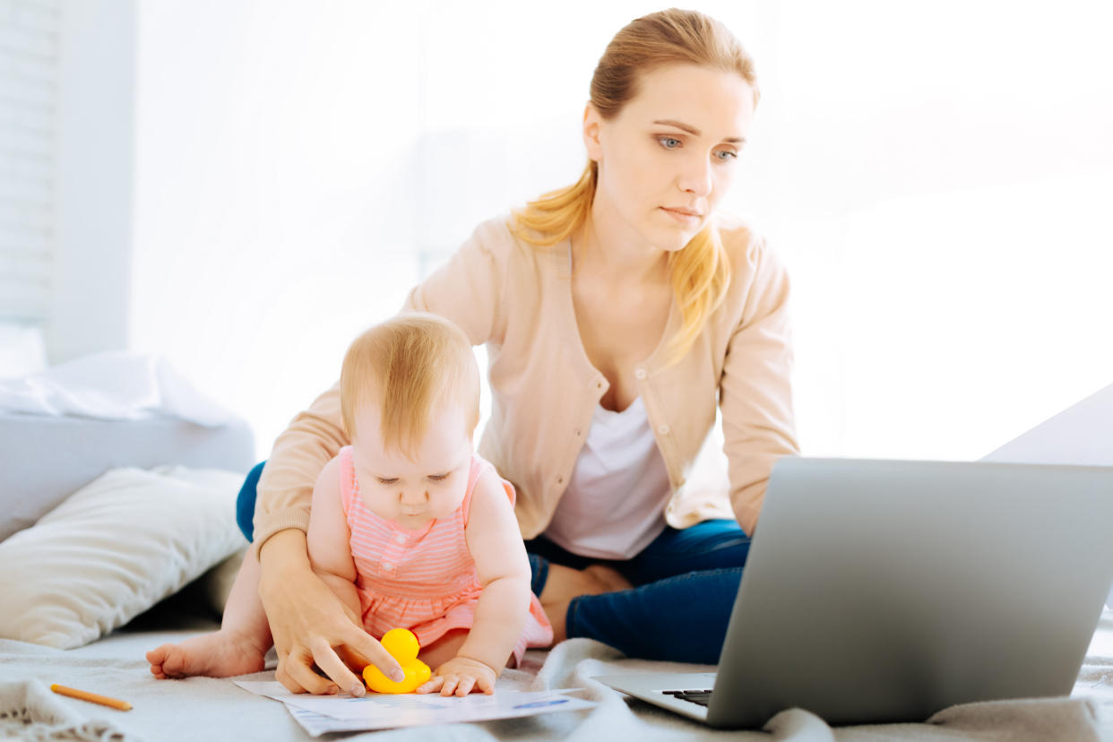 A young woman looking at her laptop while a baby sits by her side