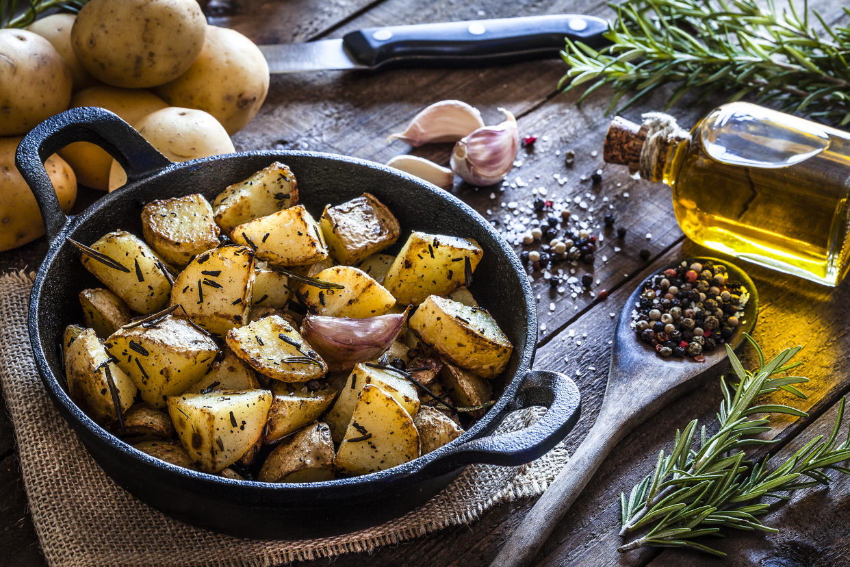 Cast iron pan filled with roasted potatoes shot on rustic wooden table. The cooking pan is at the left of an horizontal  frame and the ingredients for cooking the potatoes are all around the pan placed directly on the table. The ingredients includes are raw potatoes, rosemary, olive oil, salt, pepper and garlic. Predominant colors are brown and yellow. DSRL studio photo taken with Canon EOS 5D Mk II and Canon EF 100mm f/2.8L Macro IS USM