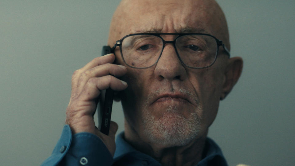 Close up of a bald, older man with a white goatee and wearing glasses. He is holding a cell phone up to his ear. He looks upset.