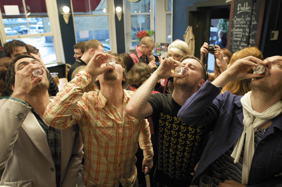 People enjoying shots at a lively indoor gathering. The setting is a casual event with a small crowd in the background. Names of individuals not provided