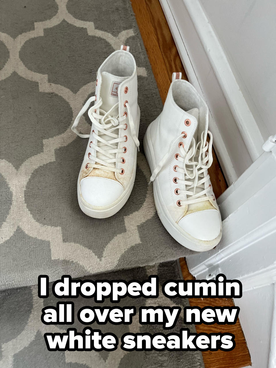 A pair of white high-top sneakers with laces are on a patterned floor next to a door