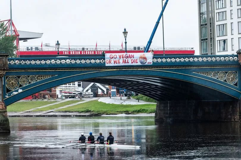 PETA supporters with the banner on Trent Bridge. The banner reads "GO VEGAN OR WE ALL DIE" written in red next to a picture of the earth on fire against a white backdrop. A group of four men in a rowing boat are seen passing underneath the banner