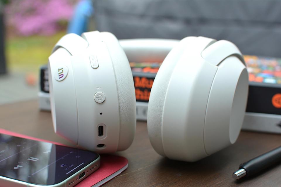 <p>White headphones sitting on a wooden table from the bottom with a red notebook and phone on the left and a black and a white book underneath.</p>
