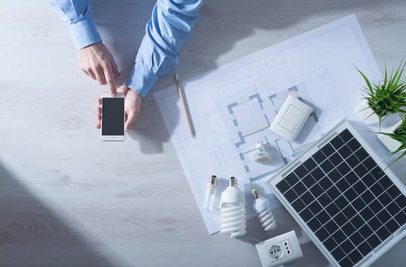 A man checking his smartphone at a desk with a small solar panel and architectural drawings sitting on it.